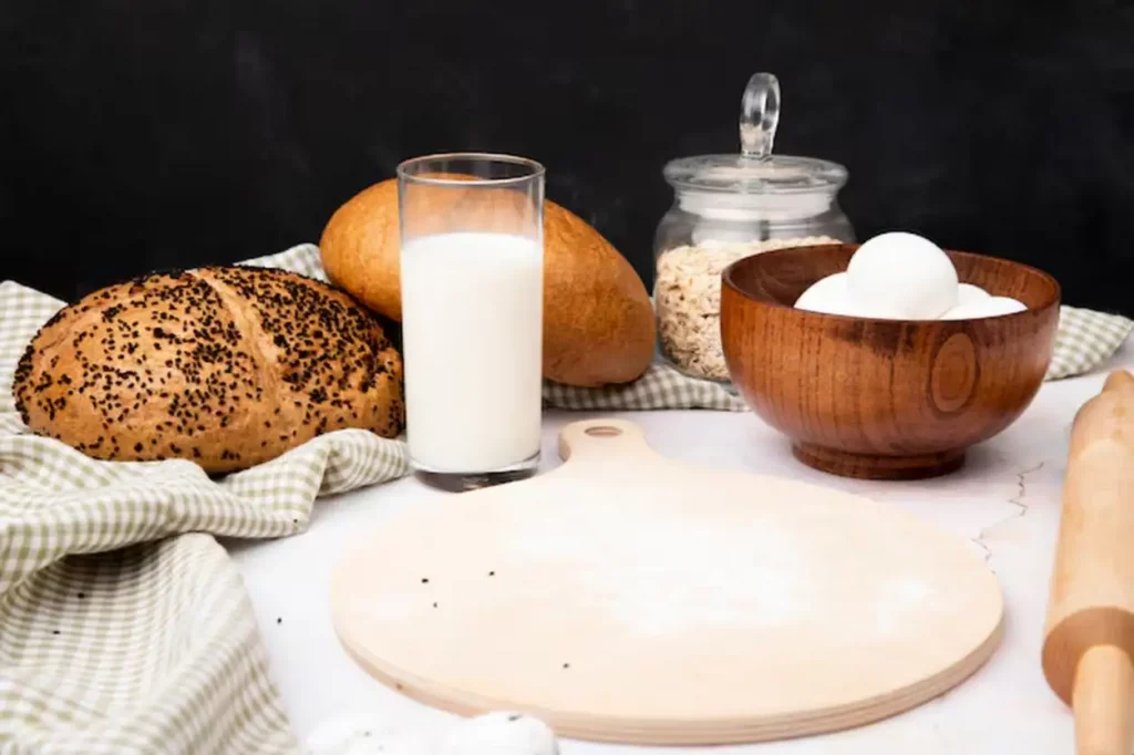 Side view of glass of milk with bowl of eggs and cutting board with cobs and oat-flakes on white surface and black background with copy space